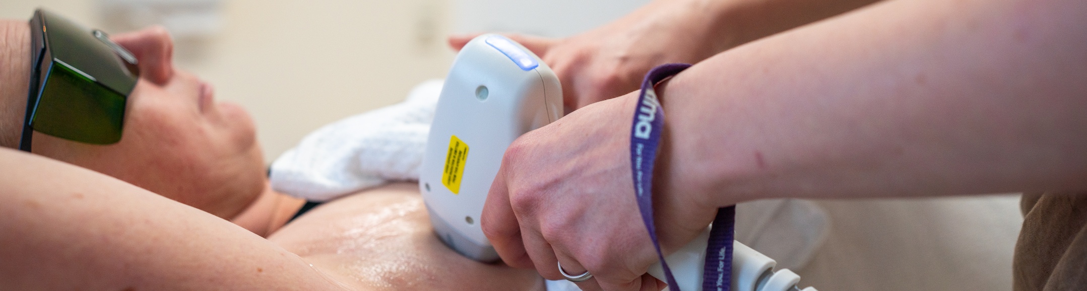 Eine Patientin erhält eine Laser-Haarentfernungsbehandlung an der Achsel. Sie trägt eine Schutzbrille, und die behandelnde Person, Eva Krüger, führt ein Handgerät über die Haut der Achsel, um die Laserenergie abzugeben.